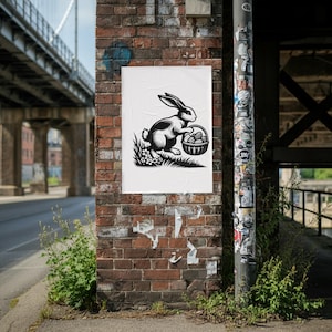 May include: Black and white illustration of a rabbit holding a basket of eggs, set against a backdrop of grass and flowers. The artwork is displayed on a white poster affixed to a brick wall.