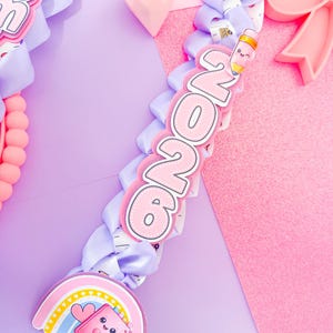 K&ouml;nnte beinhalten: Ein pastellfarbenes Abschluss-Lei mit hellvioletten B&auml;ndern und dekorativen Anh&auml;ngern. Zu den Anh&auml;ngern geh&ouml;ren ein Regenbogen, ein Notizbuch und die Zahlen "2026" in Rosa und Wei&szlig;. Der Hintergrund ist eine Mischung aus Rosa und Lila.