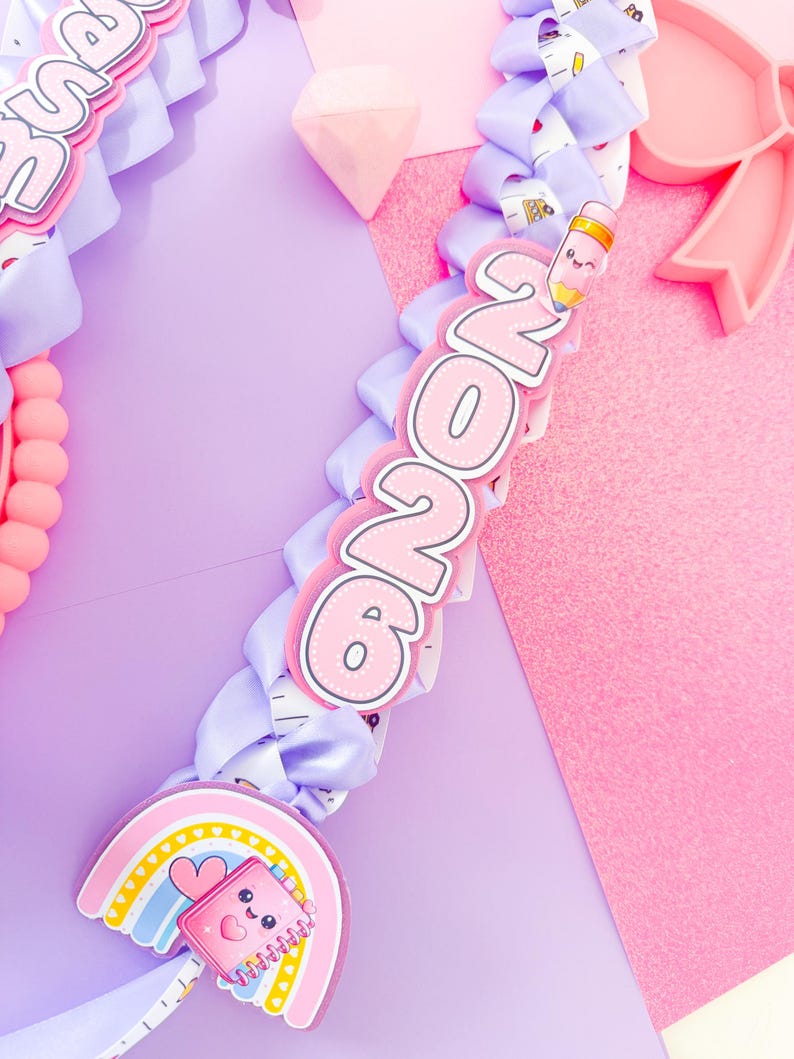 K&ouml;nnte beinhalten: Ein pastellfarbenes Abschluss-Lei mit hellvioletten B&auml;ndern und dekorativen Anh&auml;ngern. Zu den Anh&auml;ngern geh&ouml;ren ein Regenbogen, ein Notizbuch und die Zahlen "2026" in Rosa und Wei&szlig;. Der Hintergrund ist eine Mischung aus Rosa und Lila.