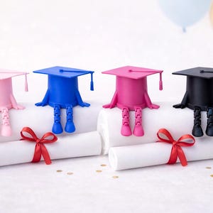 K&ouml;nnte beinhalten: Vier Dekorationen zum Thema Abschluss. Jede hat einen Mortarboard-Hut in Rosa, Blau, Rosa und Schwarz mit angebrachten Beinen. Sie sitzen auf gerollten Diplomen, die mit roten B&auml;ndern gebunden sind.
