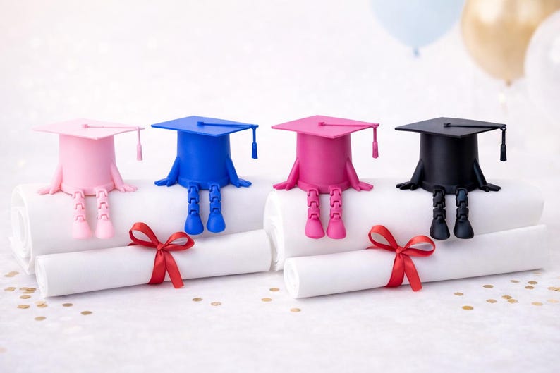 K&ouml;nnte beinhalten: Vier Dekorationen zum Thema Abschluss. Jede hat einen Mortarboard-Hut in Rosa, Blau, Rosa und Schwarz mit angebrachten Beinen. Sie sitzen auf gerollten Diplomen, die mit roten B&auml;ndern gebunden sind.