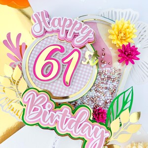 May include: Happy 61st Birthday cake topper with pink, gold, and green accents. The topper features a pink and white gingham circle with gold glitter and the number 61 in pink with gold outline. The words "Happy" and "Birthday" are in pink with gold outline and green accents. The topper is decorated with flowers and leaves.