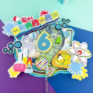 Può includere: Un colorato cake topper a tema scientifico con il numero 6. Il topper presenta varie grafiche legate alla scienza, tra cui beute, atomi ed elementi della tavola periodica. La tavolozza dei colori include blu, giallo, rosa e verde, con accenti glitterati.