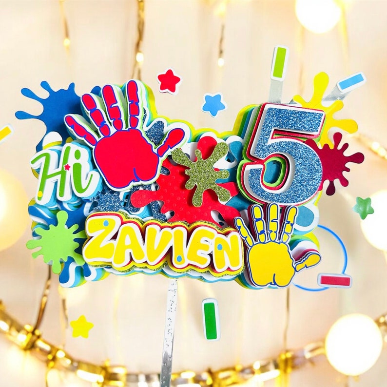 May include: A colorful cake topper with the name "ZAVIEN" and the number "5" in blue glitter. The topper also features handprints, paint splatters, and the word "Hi" in a playful font.