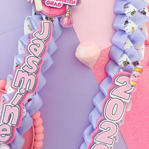 K&ouml;nnte beinhalten: Zwei Abschluss-Leis mit rosa und lila B&auml;ndern. Einer der Leis zeigt den Namen "Jamie" und eine Kindergarten-Abschlusskappe. Der andere zeigt das Jahr "2026" mit einer Bleistiftgrafik. Der Hintergrund ist eine Mischung aus Rosa und Lila.