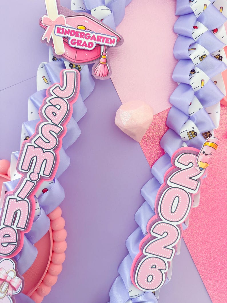K&ouml;nnte beinhalten: Zwei Abschluss-Leis mit rosa und lila B&auml;ndern. Einer der Leis zeigt den Namen "Jamie" und eine Kindergarten-Abschlusskappe. Der andere zeigt das Jahr "2026" mit einer Bleistiftgrafik. Der Hintergrund ist eine Mischung aus Rosa und Lila.