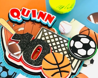 May include: A colorful sports-themed cake topper with the name "QUINN" and the number "10" in black glitter. The topper features various sports balls, including a basketball, soccer ball, baseball, and tennis ball.
