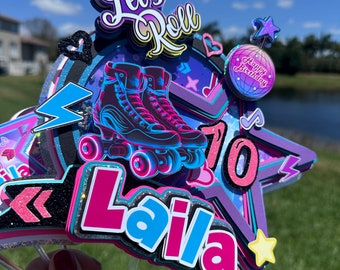 May include: A colourful cake topper featuring a star, roller skates, and the text "Let's Roll" and "Laila 10". The topper is decorated with glitter and has a rainbow colour scheme.