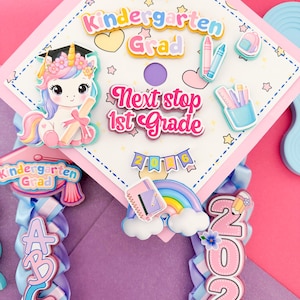 Puede incluir: Un birrete de graduación de jardín de infancia rosa y blanco decorado con un unicornio, libros, crayones y el texto "Kindergarten Grad" y "Next stop 1st Grade". El birrete tiene una cinta azul y morada con las letras A, B, C, D y el año 2026.