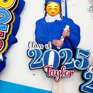 Puede incluir: Un accesorio fotográfico con temática de graduación con birrete y toga azules, con el texto "Class of 2025 Taylor". El accesorio incluye un mango de palo de madera y una combinación de colores azul y blanco. Accesorios adicionales: un búho y un cuaderno.