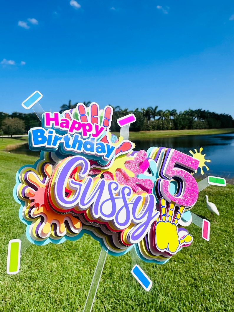 May include: A colorful cake topper with the words "Happy Birthday" and "Gussy" in purple, pink, and yellow. The number "5" is in pink with glitter and a yellow handprint. The topper is decorated with colorful paint splatters.