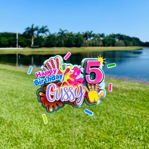 May include: A colorful cake topper with the words "Happy Birthday" and the name "Gussy" in pink and purple lettering. The number "5" is in pink with a yellow sun and handprint design. The topper is decorated with colorful paint splatters and handprints.