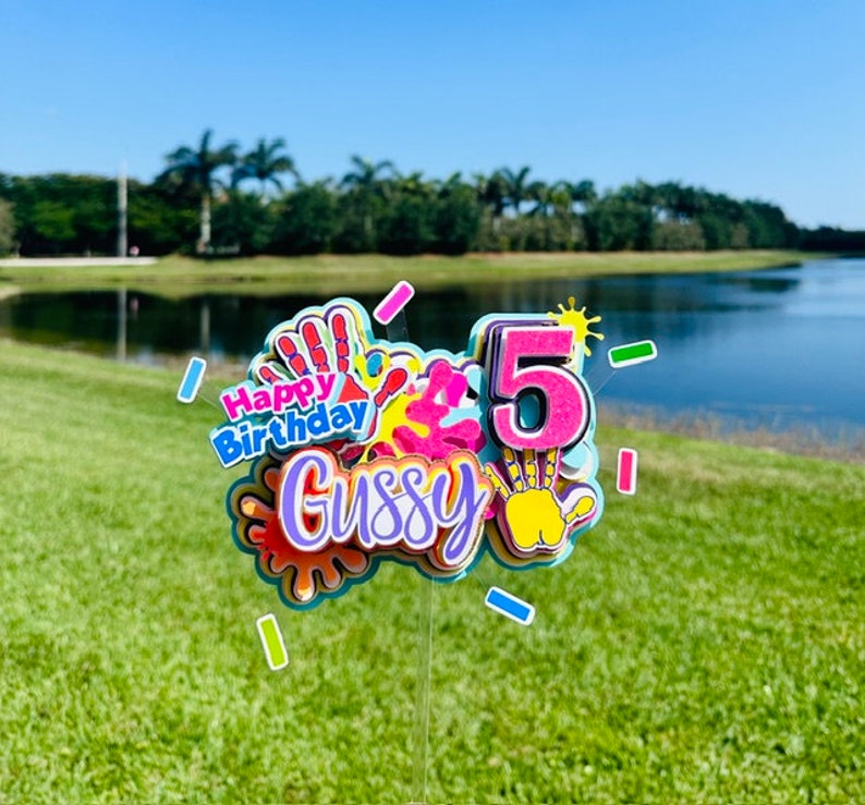 May include: A colorful cake topper with the words "Happy Birthday" and the name "Gussy" in pink and purple lettering. The number "5" is in pink with a yellow sun and handprint design. The topper is decorated with colorful paint splatters and handprints.
