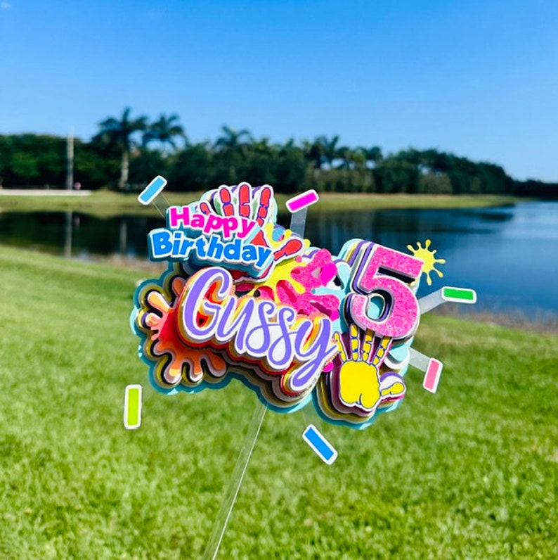 May include: A colorful cake topper with the words "Happy Birthday" and the name "Gussy" in a rainbow font. The number "5" is also included in the topper, with a glittery finish. The topper is decorated with handprint designs and colorful paint splatters.