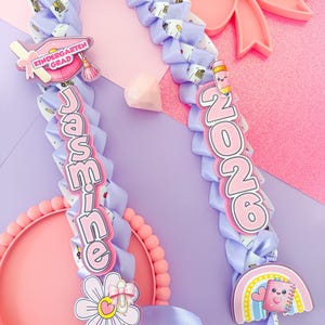 K&ouml;nnte beinhalten: Eine lila Abschluss-Lei mit dem Namen "Jasmine" und dem Jahr "2026" in rosa Schrift. Die Lei ist mit schulischen Charms verziert, darunter eine Abschlusskappe, ein Bleistift und ein Regenbogen-Notizbuch. Eine rosa Schleife und ein Teller sind im Hintergrund.