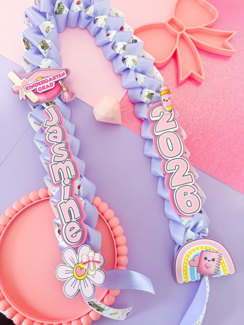 K&ouml;nnte beinhalten: Eine lila Abschluss-Lei mit dem Namen "Jasmine" und dem Jahr "2026" in rosa Schrift. Die Lei ist mit schulischen Charms verziert, darunter eine Abschlusskappe, ein Bleistift und ein Regenbogen-Notizbuch. Eine rosa Schleife und ein Teller sind im Hintergrund.