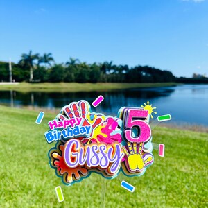 May include: A colorful cake topper with the words "Happy Birthday" and the name "Gussy" in a rainbow font. The topper also features a number "5" with glitter and a handprint design.