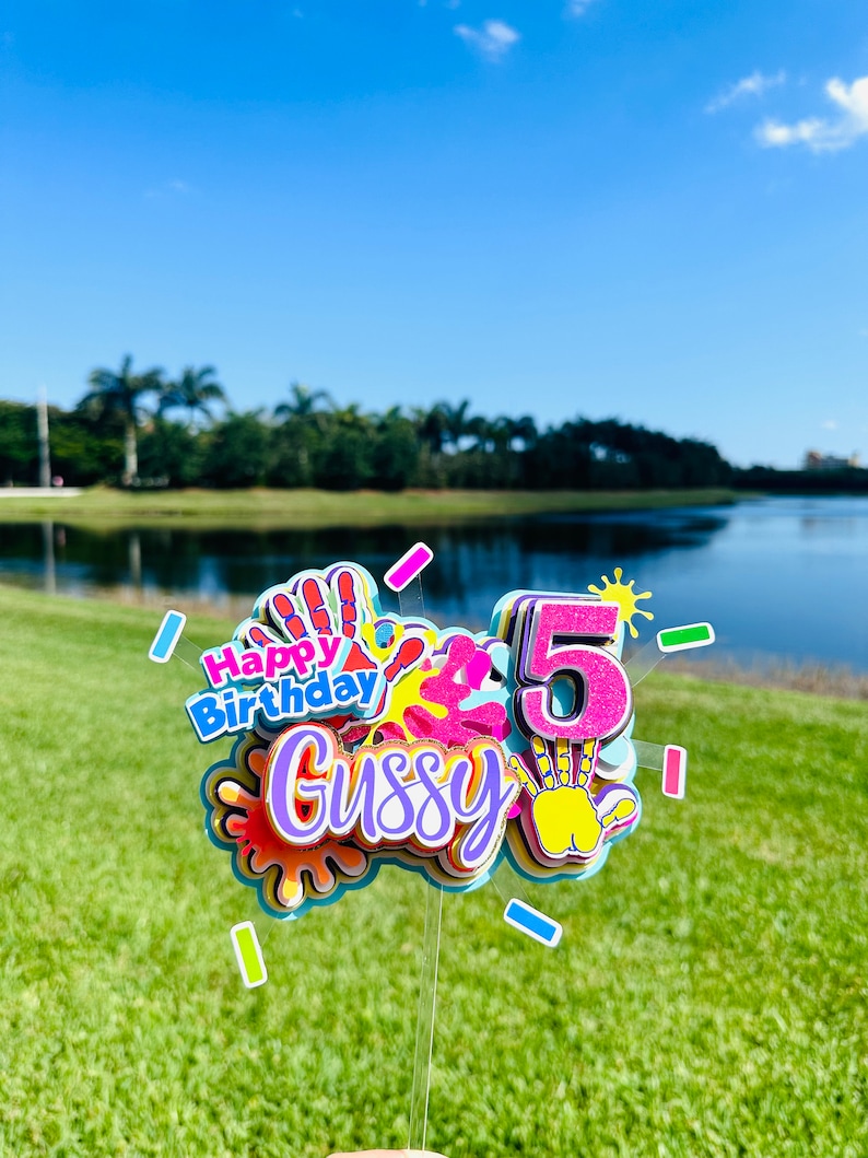 May include: A colorful cake topper with the words "Happy Birthday" and the name "Gussy" in a rainbow font. The topper also features a number "5" with glitter and a handprint design.