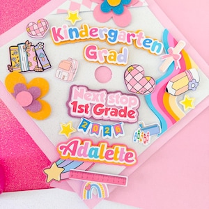 Puede incluir: Un gorro de graduación rosa y blanco decorado con adornos coloridos. El gorro presenta el texto "Kindergarten Grad", "Next stop 1st Grade" y el nombre "Adalette". Los elementos decorativos incluyen flores, estrellas, corazones, un lápiz y un arcoíris.