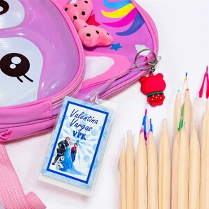 May include: A pink unicorn backpack with a white and blue Frozen character name tag that says "Valentina Vargas VPK". The tag is attached to a clear plastic strap with a red strawberry keychain. There are also wooden dowels with colorful tips.