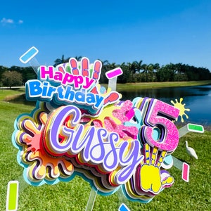 May include: A colorful cake topper with the words "Happy Birthday" and "Gussy" in purple, pink, and yellow. The number "5" is in pink with glitter and a yellow handprint. The topper is decorated with colorful paint splatters.