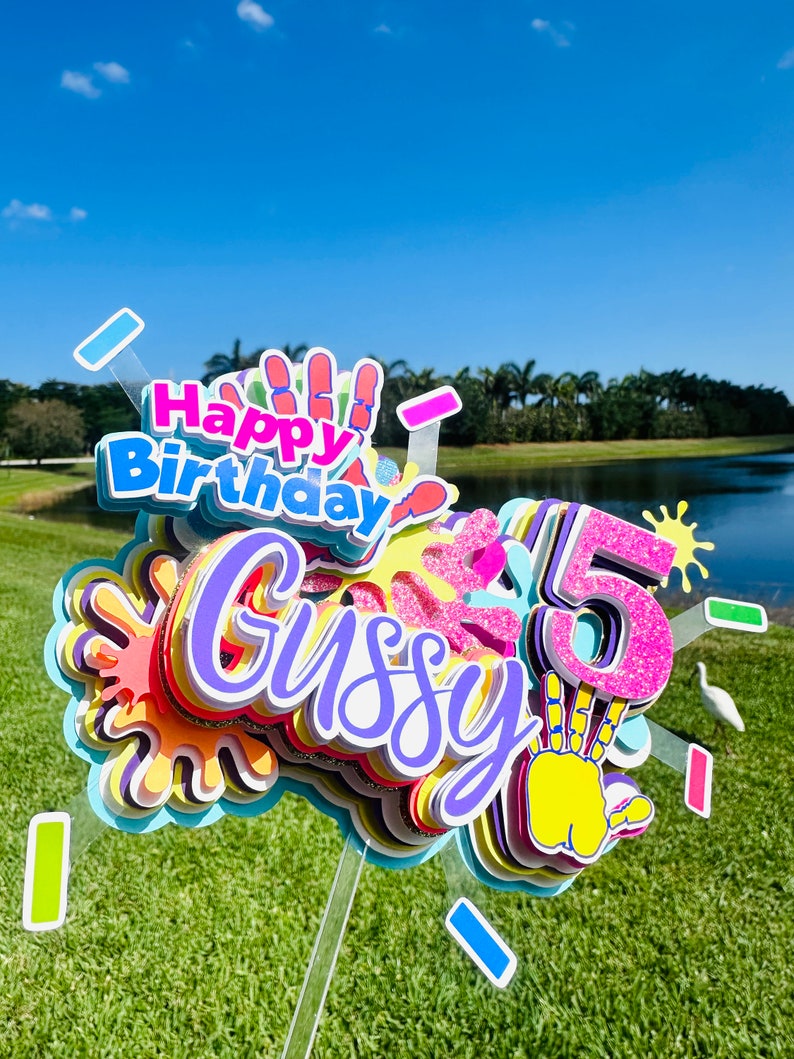 May include: A colorful cake topper with the words "Happy Birthday" and "Gussy" in purple, pink, and yellow. The number "5" is in pink with glitter and a yellow handprint. The topper is decorated with colorful paint splatters.