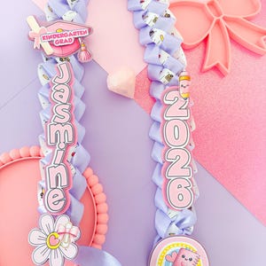 Puede incluir: Un lei de cinta lavanda con el nombre "Jasmine" y el año "2026" en letras rosas. El lei está decorado con amuletos temáticos de graduación, incluyendo un birrete, un arcoíris y una flor. El fondo es una mezcla de rosa y morado.