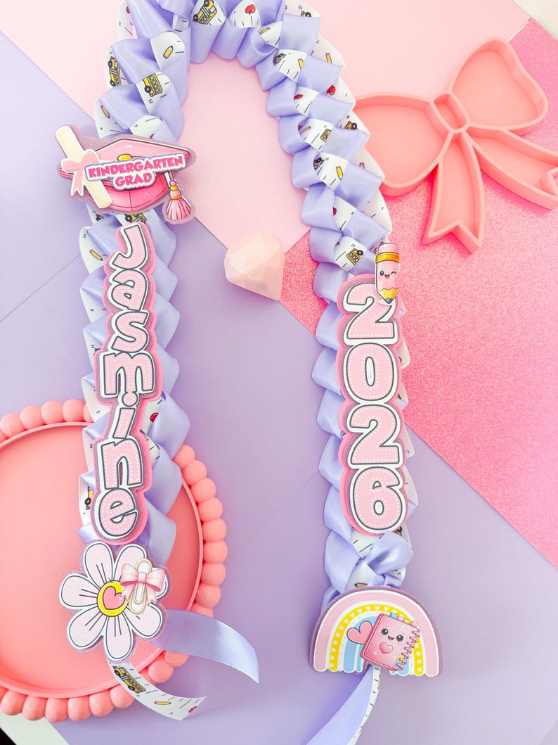 K&ouml;nnte beinhalten: Ein lavendelfarbenes Band-Lei mit dem Namen "Jasmine" und dem Jahr "2026" in rosa Schrift. Das Lei ist mit Abschluss-Themen-Anh&auml;ngern verziert, darunter ein Hut, ein Regenbogen und eine Blume. Der Hintergrund ist eine Mischung aus Rosa und Lila.