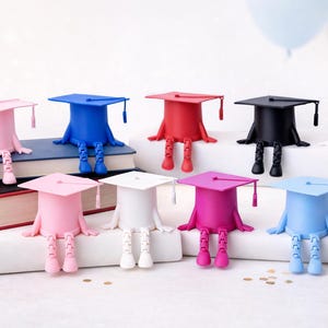K&ouml;nnte beinhalten: Eine Sammlung farbenfroher Abschlusskappen-Figuren in Rosa, Blau, Rot, Schwarz und Wei&szlig;. Jede Kappe hat eine Quaste und sitzt auf einem Buch oder einer wei&szlig;en Oberfl&auml;che, mit herunterh&auml;ngenden Beinen. Ballons sind im Hintergrund.