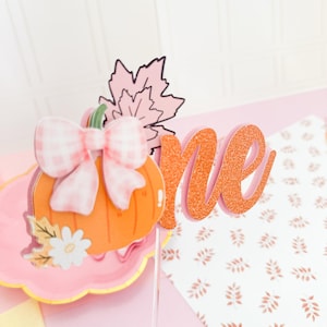 May include: A festive orange pumpkin cake topper with a pink gingham bow, a pink leaf, and the word "one" in glittery orange. The topper sits on a pink plate with a floral design, perfect for a first birthday celebration.