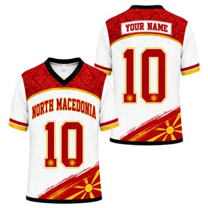 May include: A white V-neck sports jersey featuring red and yellow accents. The front displays the text NORTH MACEDONIA above the number 10. The back shows the text YOUR NAME above the number 10. Both sides feature a sunburst graphic pattern.