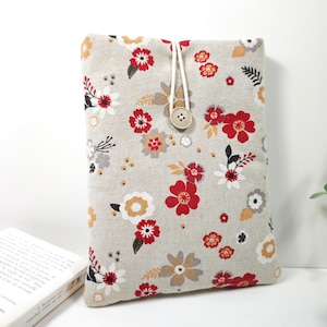 Peut inclure: Une housse de livre en lin beige avec un motif floral rouge, blanc et jaune. La housse a une fermeture à bouton et un cordon de serrage blanc.