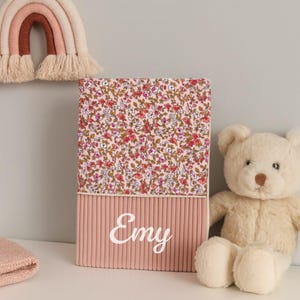 Puede incluir: Una caja de almacenamiento rosa personalizada con el nombre "Emy" en escritura blanca. La sección superior presenta un estampado floral con flores rojas y rosas, mientras que la inferior tiene una textura acanalada. Un oso de peluche beige y un arcoíris colgante de pared están en el fondo.