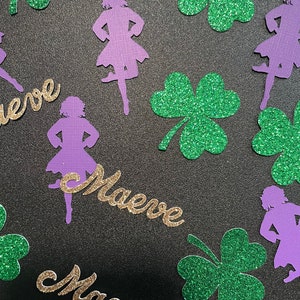 May include: Purple glitter paper cutouts of a girl in a dancing pose with the name "Maeve" written in gold glitter paper. Green glitter paper shamrocks are scattered around the cutouts.