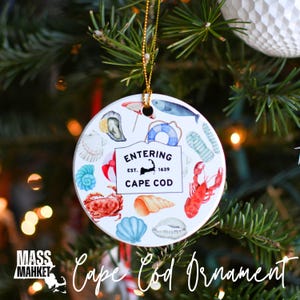 May include: Round ceramic Christmas ornament with a Cape Cod theme. The ornament features illustrations of seashells, a lobster, and a sign that reads "Entering Cape Cod". The ornament is hanging on a Christmas tree.