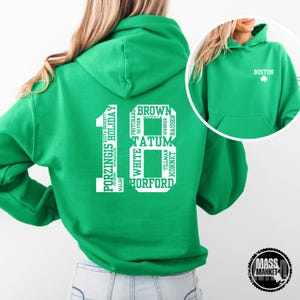Może przedstawiać: Zielony sweter z kapturem z białą liczbą 18 z tyłu. Liczba składa się z nazwisk graczy Boston Celtics. Z przodu swetra znajduje się napis "BOSTON" białymi literami z koniczyną.
