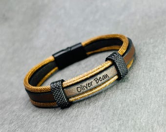 May include: A brown and black leather bracelet with a gold accent and the name "Oliver Bean" engraved on it.