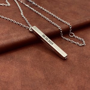Può includere: Una collana con ciondolo a forma di barra in argento. La barra è incisa con la data 06.06.2024.