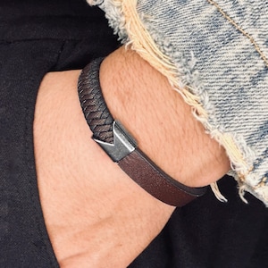 May include: A brown leather bracelet featuring a woven section and a silver-toned clasp. The bracelet is worn on a wrist, with a denim jacket sleeve visible. A fashionable accessory for everyday wear.