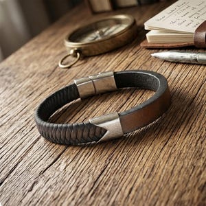 Könnte beinhalten: Ein braunes und schwarzes Lederarmband mit Silberverschluss, auf einer Holzoberfläche. Das Armband hat ein geflochtenes Design auf dem schwarzen Lederteil und ein glattes braunes Lederband. Ein Kompass und ein Notizbuch sind im Hintergrund.