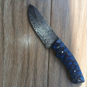 May include: A Damascus steel knife with a patterned blade and a blue and black handle. The handle has a geometric design with white accents. The knife is set against a wooden background.