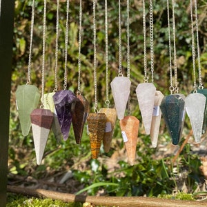 May include: A collection of 14 crystal pendulums hanging from silver chains. The pendulums are made of various types of crystals, including amethyst, tiger's eye, rose quartz, and green aventurine.
