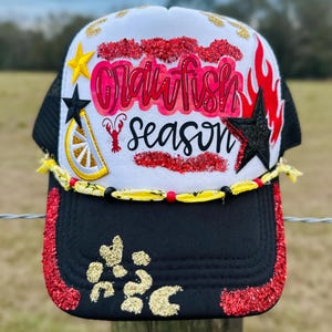 May include: A trucker hat with a black mesh back and a white front. The front features the text "Crawfish Season" in pink and red, along with a crawfish illustration and star accents. The brim is black with red glitter.