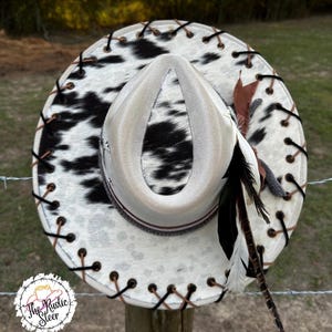 May include: Cowboy hat with a white and black cowhide pattern on the brim. The hat has a cream-coloured crown, brown and black stitching, and decorative feathers. The Rustic Steer logo is visible on the bottom left.