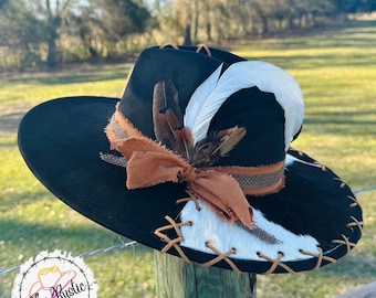 May include: A black felt cowboy hat with a white and brown cowhide band, brown leather lacing, and a brown and white feather accent. The hat is sitting on a wooden fence post.