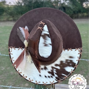 Könnte beinhalten: Ein brauner Cowboyhut mit einem weißen und braunen Kuhfellband. Der Hut hat eine braune Wildlederkrone und eine weiße Kuhfellkrempe. Der Hut ist mit einer braunen Feder und einem braunen Lederriemen verziert.