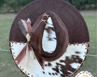 May include: A brown cowboy hat with a white and brown cowhide band. The hat has a brown suede crown and a white cowhide brim. The hat is decorated with a brown feather and a brown leather strap.