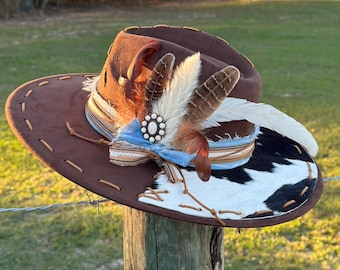May include: A brown cowboy hat with a wide brim, adorned with feathers, a blue ribbon, and a decorative centerpiece. The hat features a cowhide patch and stitched detailing, creating a rustic and stylish accessory.