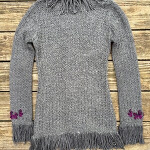 May include: A gray knit sweater with long sleeves and fringe detailing. The sweater features fringe around the collar, cuffs, and hem. Small purple floral embellishments are on the cuffs. The sweater is made of a soft, textured knit fabric.