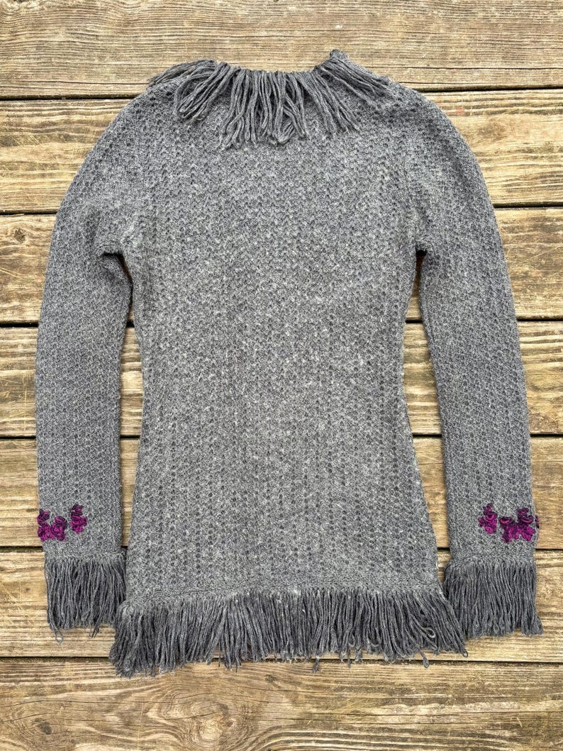 May include: A gray knit sweater with long sleeves and fringe detailing. The sweater features fringe around the collar, cuffs, and hem. Small purple floral embellishments are on the cuffs. The sweater is made of a soft, textured knit fabric.